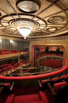 an empty theater with red seats and chandelier above the stage, looking down at the auditorium