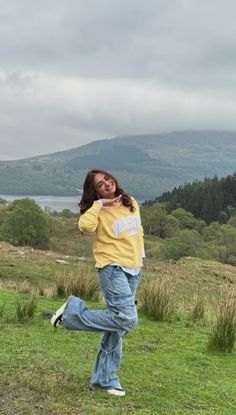 a woman in yellow shirt and jeans standing on grass with mountains in the back ground