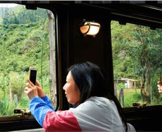 two women are taking pictures on their cell phones while traveling in a train with mountains and trees behind them