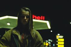 a man with long hair standing in front of a gas station at night, looking off to the side