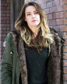 a woman leaning against a brick wall wearing a green jacket and fur collared coat