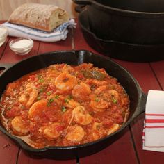 a skillet filled with shrimp and sauce on top of a wooden table next to a loaf of bread