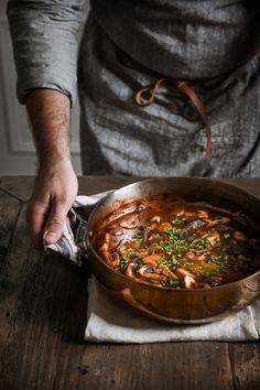 a person holding a pan filled with food on top of a wooden table next to a knife