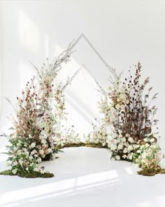 an arrangement of flowers and greenery in front of a white wall with a mirror