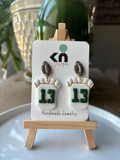 a pair of football themed earrings is displayed on a small easel with a plant in the background