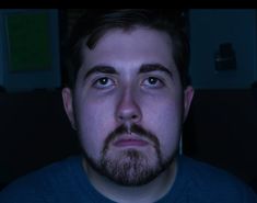 a man with a beard and blue eyes stares into the camera while sitting in a dark room