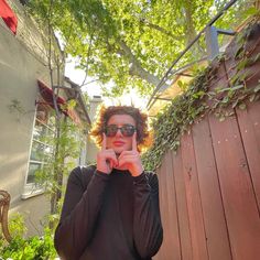 a woman with red hair and sunglasses is holding her hands to her face while standing in front of a wooden fence