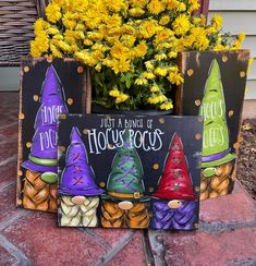 three painted wooden signs sitting on top of a brick floor next to yellow flowers and a potted plant