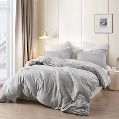 a bed with grey sheets and pillows in a white room next to a large window