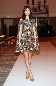 a woman is walking down the runway wearing a dress with flowers on it and brown shoes