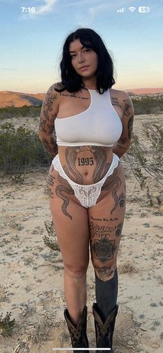 a woman with tattoos on her body and cowboy boots standing in the desert, posing for a photo