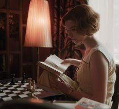 a woman sitting at a table with a book in her hands and chess on the table