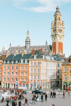 many people are walking around in front of some buildings and a clock tower on the other side