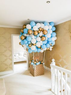 a teddy bear in a basket with blue and white balloons floating from the ceiling above