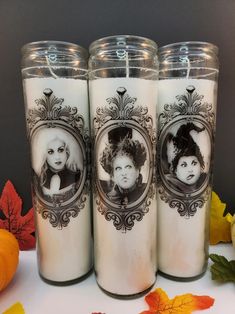 three white candles with black pictures on them and autumn leaves in the foreground, along with two pumpkins