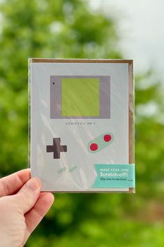 a hand holding up a gameboy card in front of some trees and grass,