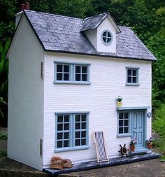 a white doll house sitting on top of a wooden platform in front of trees and bushes