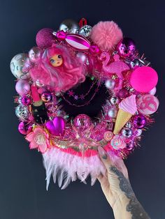 a hand holding up a pink wreath decorated with barbie dolls and other accessories on it