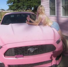 a blonde woman leaning on the hood of a pink mustang parked in front of a house