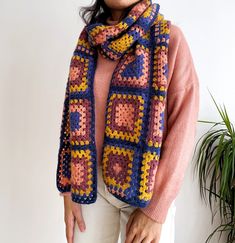 a woman wearing a crocheted scarf and pink sweater stands in front of a potted plant