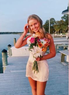 a beautiful young woman holding a bouquet of flowers while standing on a dock next to the water