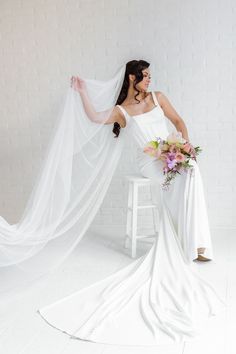 A Bride Sitting On A Chair Holding Her Veil And Holding Her Bridal Bouquet Looking Off To The Side.