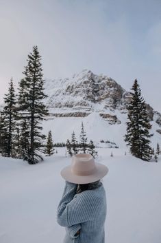 a woman wearing a hat standing in the snow with her arms behind her head looking at a mountain