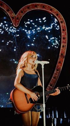 a woman holding a guitar while standing in front of a heart shaped stage with fireworks behind her