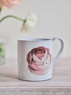 a coffee mug with a pink rose painted on the front and side, sitting on a table next to a flower vase