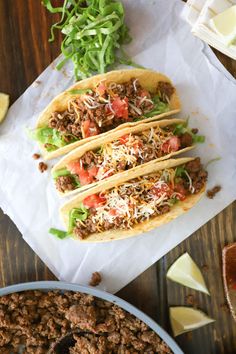 three tacos sitting on top of a table next to a bowl of salsa and lime wedges