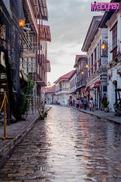 a wet street with people walking on it and buildings in the background at sunset or dawn