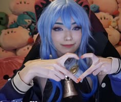 a woman with blue hair making a heart sign while sitting in front of stuffed animals