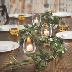 a wooden table topped with lots of white plates and glasses filled with candles surrounded by greenery