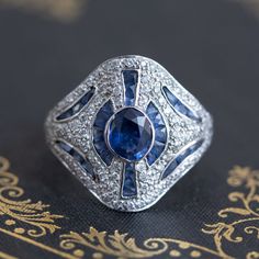 A fun and unusual cocktail ring, the styling of this ring is reminiscent of Art Deco themes with the geometric lines and the use of diamonds alongside blue sapphires. The central stone is a faceted medium blue oval sapphire of approximately 1.00ct, surrounded by 28 calibre cut sapphires (approx. .75ctw); there are also .50ctw of diamonds added in for good measure to yield an ornate yet subtly fancy ring! The setting is low-profile making it very easy and comfortable to wear. 18kt white gold; siz Art Deco Oval Diamond Ring With Accent Stones, Art Deco Blue Diamond Ring, Blue Diamond Art Deco Ring, Blue Art Deco Diamond Ring, Blue Ring With Single Cut Diamonds, Fine Jewelry, Fine Jewelry Blue Rings With Single Cut Diamonds, Art Deco Blue Diamond Ring With Accents, Art Deco Blue Diamond Ring With Center Stone, Blue Art Deco Diamond Ring With Center Stone