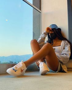 a woman sitting on the floor next to a window with her legs crossed and wearing white sneakers