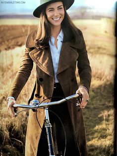 a woman wearing a hat and coat riding a bike in a field with dry grass