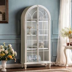 a white china cabinet with glass doors and flowers in vases on the floor next to it