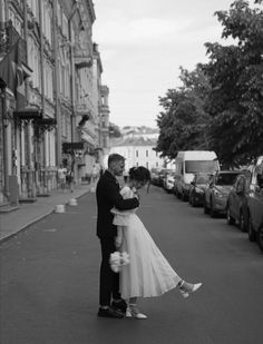 a bride and groom kissing on the street in front of some parked cars, black and white photograph