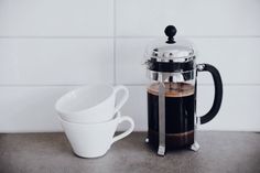 two coffee cups sitting next to each other on top of a counter with a french press