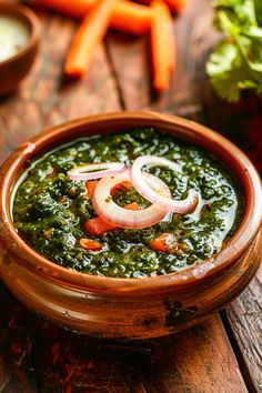 a wooden bowl filled with green soup and onions on top of a table next to carrots