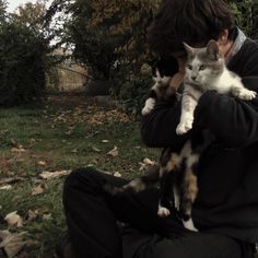 a man holding a cat in his arms while sitting on top of a grass covered field