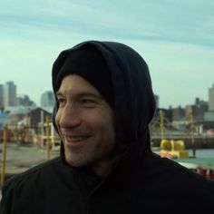 a man wearing a black hoodie smiles at the camera in front of a cityscape