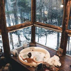 a woman is sitting in the bathtub looking out at the snow covered forest outside