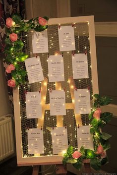 a wedding seating chart with pink roses and greenery on the side, surrounded by fairy lights