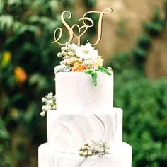 a three tiered white marble cake with flowers and initials on the top, sitting in front of greenery