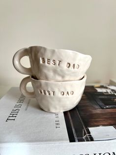 two white ceramic cups sitting on top of a stack of books with the words dad test and best dad printed on them