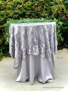 a table covered with a silver cloth and flowers in front of shrubbery on the sidewalk