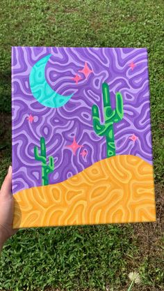 a hand holding up a purple and green painting with cactuses in the desert on it