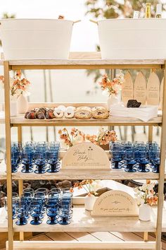 an outdoor dessert table with blue glasses and flowers on the top shelf, along with other food items