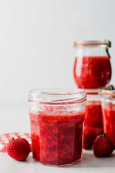 three jars filled with strawberry jam on top of a white table next to strawberries